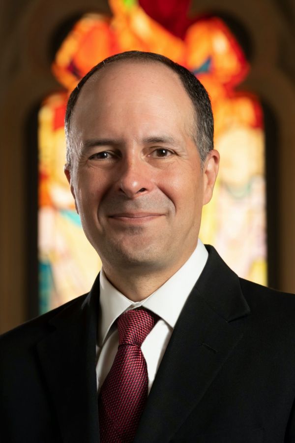 Edgardo Colón-Emeric headshot in black suit and red tie in front of Pentecost window