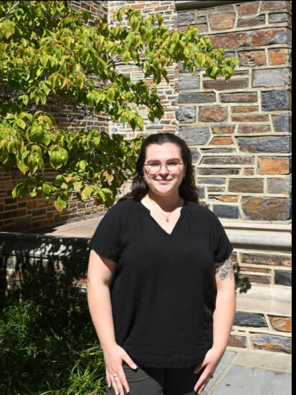 Maggie Kennedy in black shirt and dark green pants in front of Divinity brick