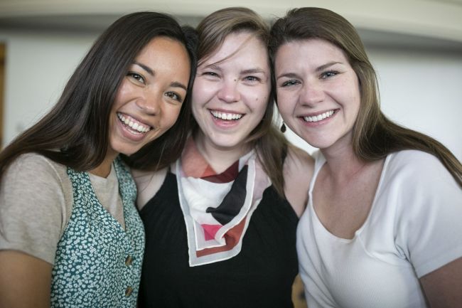 Three female students pose smiling, (far left) in green floral shirt, (middle) in black shirt and colorful scarf, (far right) white shirt