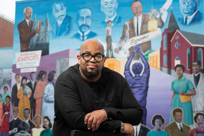 Eric Williams wearing black clothing and glasses sits in front of Durham Civil Rights mural with his arms folded