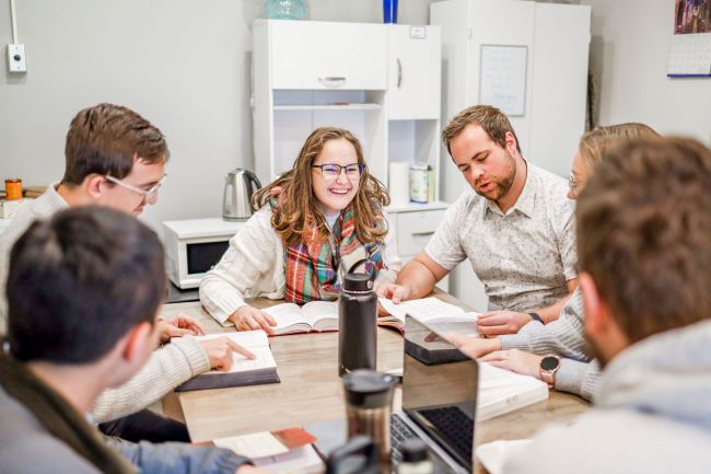 Six AEHS students studying around a table: featuring a woman in a tan sweater and plaid scarf, a man with a beard and a white short sleeve button down shirt, and others