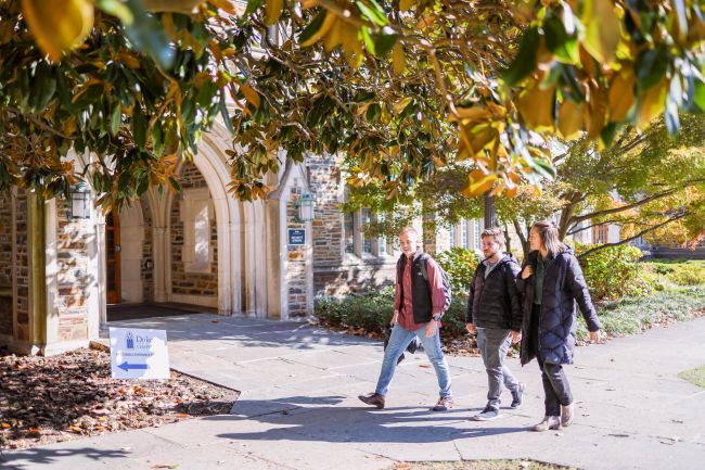 Three students walking outside past the Divinity School: woman in green shirt, man with dark hair and beard in a black coat, and a man with light hair in a red shirt and gray vest