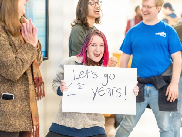 A student with pink hair holds a sign that reads "Let's go first years!"