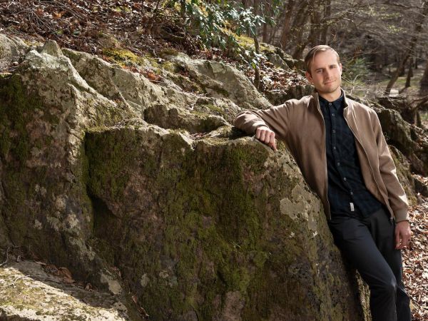 Alum Avery Davis Lamb poses in black shirt and tan jacket in front of rock