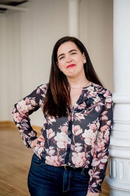 Kate Bowler is wearing a flowered shirt, with her hand on her hip