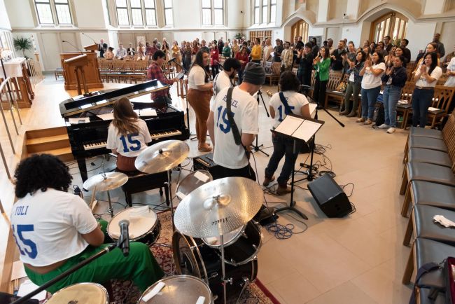 Band members and drumset from behind with people in background standing and clapping