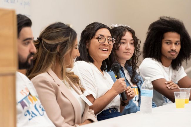 Student speaks during a panel discussion in white shirt and black rimmed circle glasses