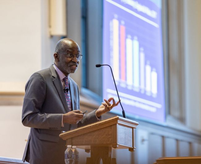 Dr. David R. Williams, wearing a suit, stands at the podium and explains a graph on a slide in the background in Goodson Chapel