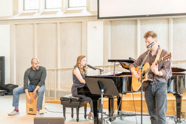 Three students play music for worship in Goodson Chapel. (far left) student in black shirt and jeans play box drum, (middle) student in black pant suit plays piano, (far right) student in brown shirt and jeans plays guitar