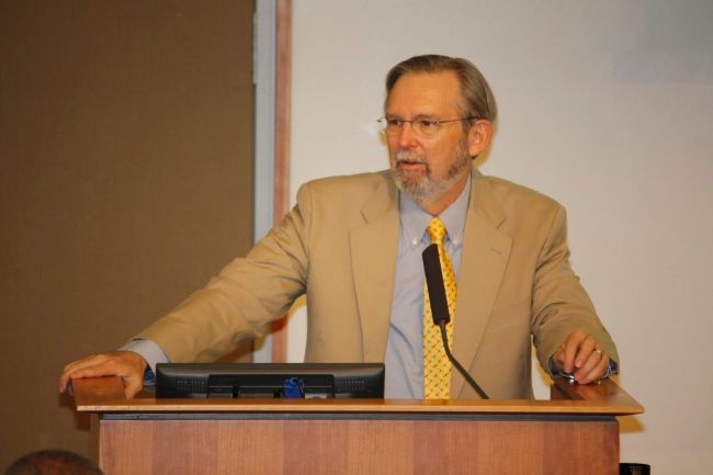 Richard stands at a classroom podium wearing tan jacket and yellow tie