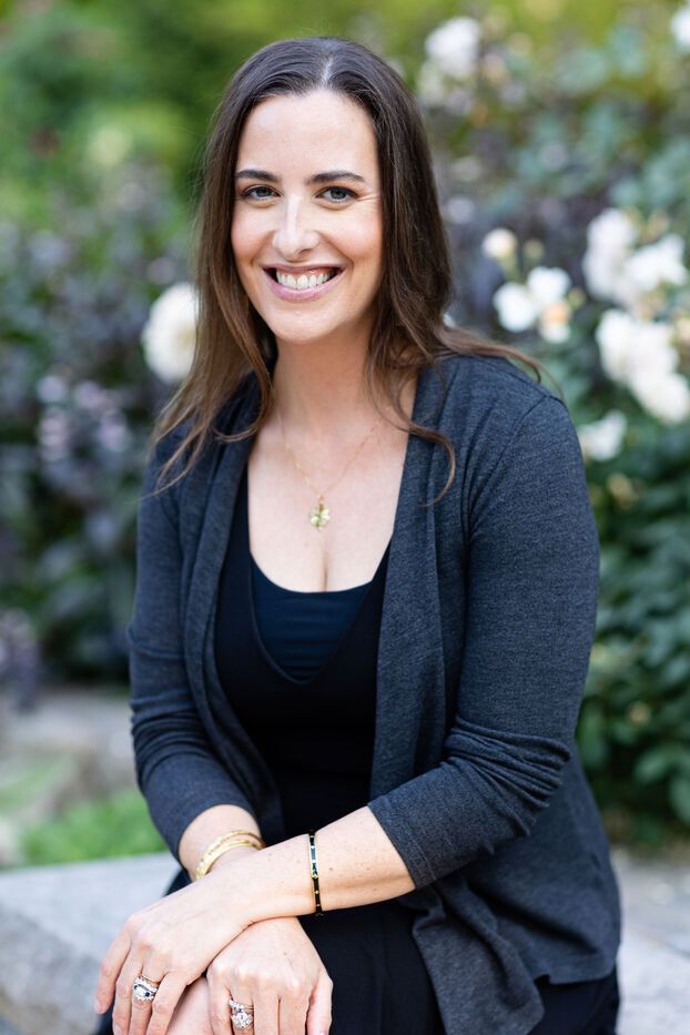 Dana Trent, a woman with light skin and dark brown hair is sitting in front of green bushed with white flowers. She is wearing a black shirt with a gray cardigan.