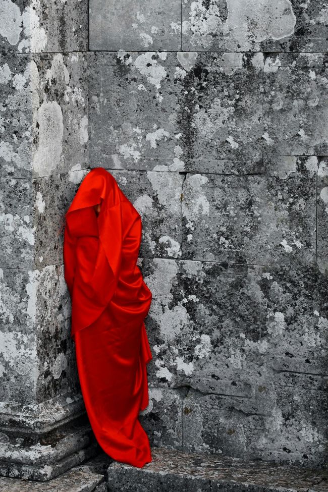 Person wrapped in red silk stands against stone wall