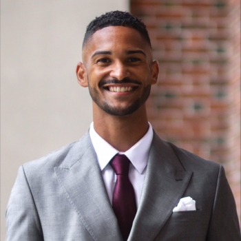 Aaron Shirley smiling wearing a grey suit with burgundy tie and holding a graduation cap