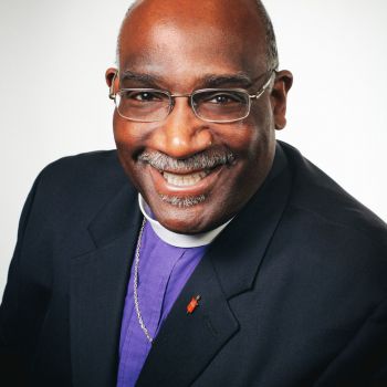 Image of a smiling man wearing glasses clerical collar, purple clerical shirt, and navy suit coat