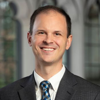 Daniel Corpening headshot in front of Divinity School brick exterior, wearing a dark blue sweater and tie with white dress shirt