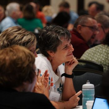 Convocation on the rural church gathering with people sitting around tables in discussion, featuring a women in a white shirt listening intently