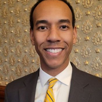 Harrison Hines smiling wearing a grey blazer, white shirt, and yellow tie