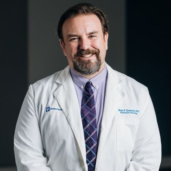 Brian Quaranta wearing a purple shirt, tie, and white medical coat