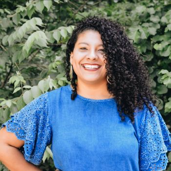 Yolanda is smiling and standing in front of plants. She is wearing a blue dress and gold earrings.