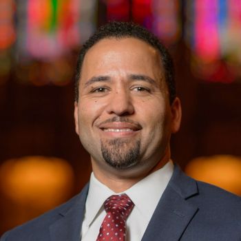 Luke Powery headshot in front of colorful Duke Chapel stained glass in dark gray suit with red tie