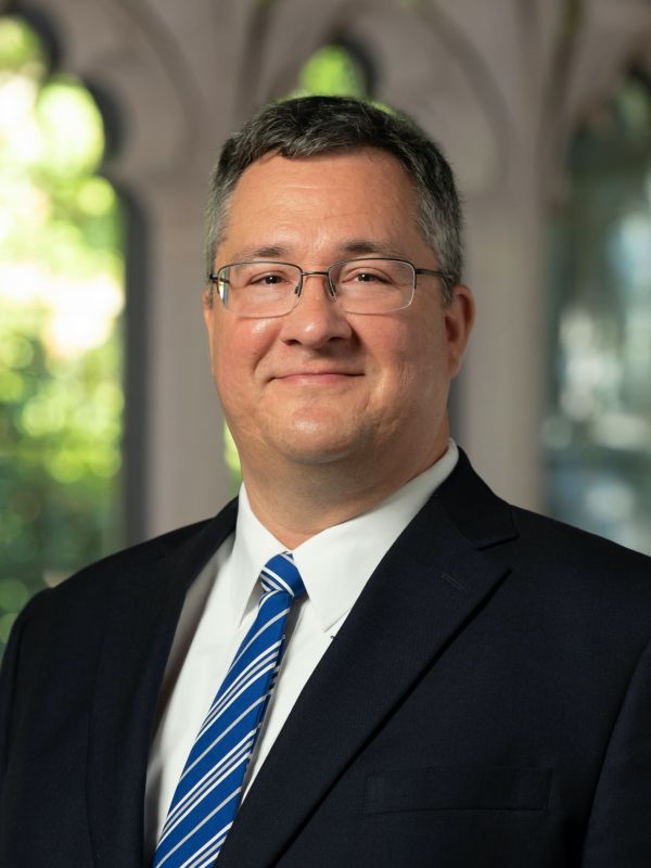 Daniel Castelo headshot in black suit and blue tie with white stripes