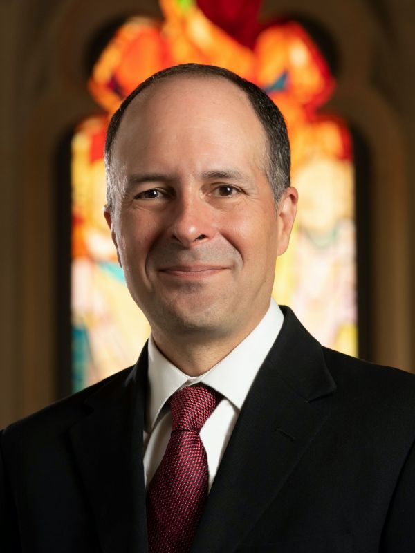 Edgardo Colón-Emeric headshot in black suit and red tie in front of Pentecost window